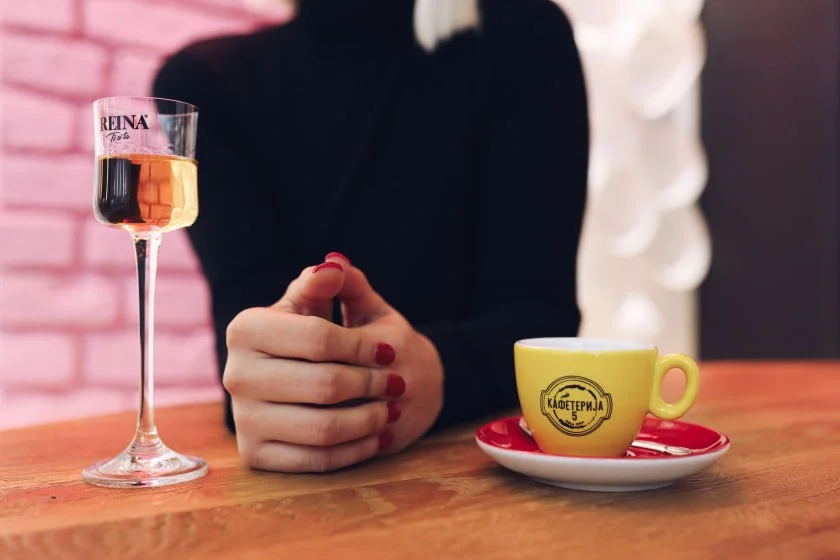 Čaša sa pićem stoji lijevo, ruke sklopljene u sredini, žuta šoljica desno na stolu. Tekst: "REINA" i "Кафетерија".