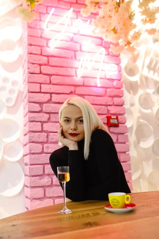 Žena sjedi, oslonjena rukom i gleda kameru, ispred nje čaša pića i šoljica, ispred rozog zida s neonskim natpisom.