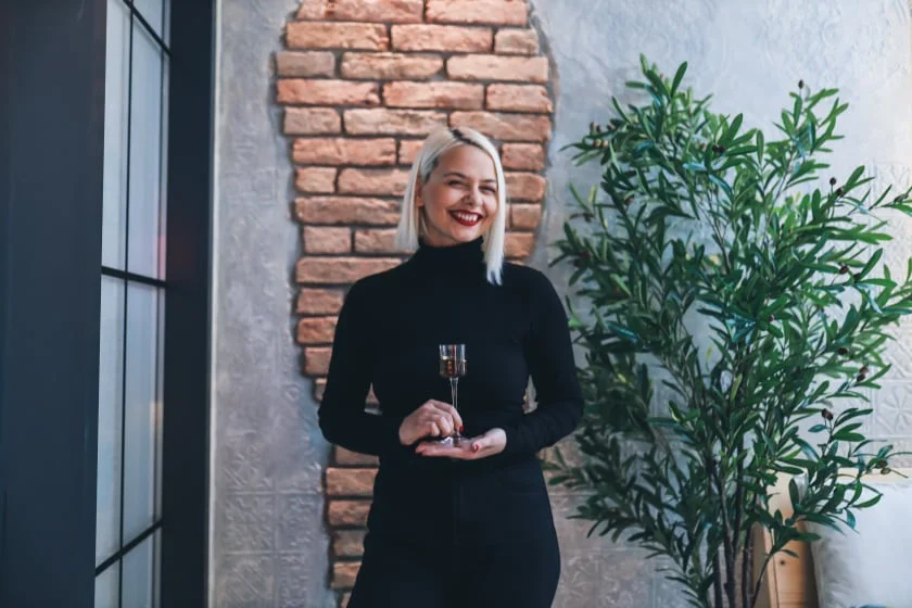 Osoba drži čašu, nasmijana je, stoji ispred zida s ciglama i biljkom u zatvorenom prostoru.