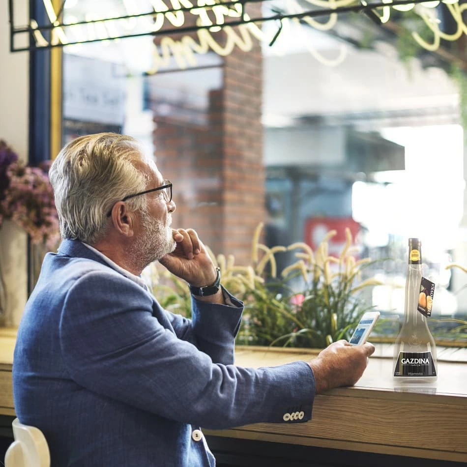 Muškarac sjedi, drži telefon, pored boce "GAZDINA" likera na stolu u kafiću.