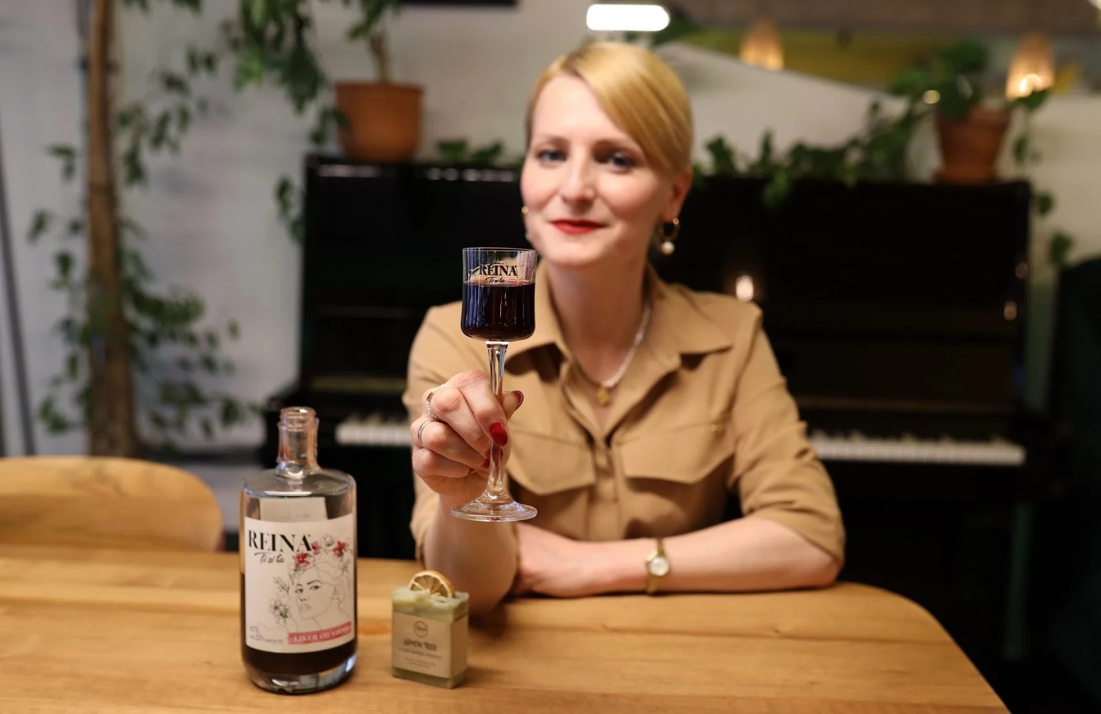 Osoba drži čašu vina na stolu u sobi sa klavirom u pozadini. Na boci piše "REINA".