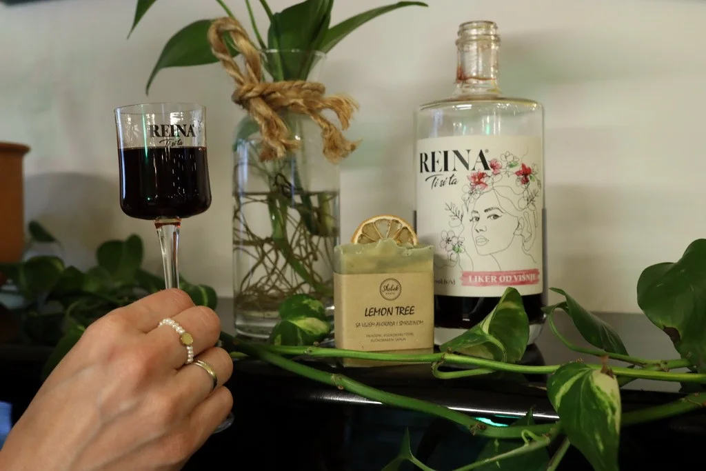 Čaša vina u ruci, boca likera i sapun, smješteni na stolu sa zelenilom. Tekst: "REINA Ti si ta LIKER OD VIŠNJE" i "LEMON TREE".