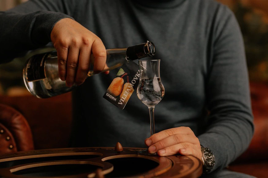 Osoba sipa piće iz boce u čašu na stolu. Tekst: "GAZDIĆ", "VILIJAMOVKA".