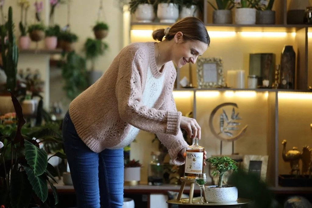 Osoba stavlja bocu na sto u sobi sa biljkama i ukrasima. Tekst na boci: "REFINA".
