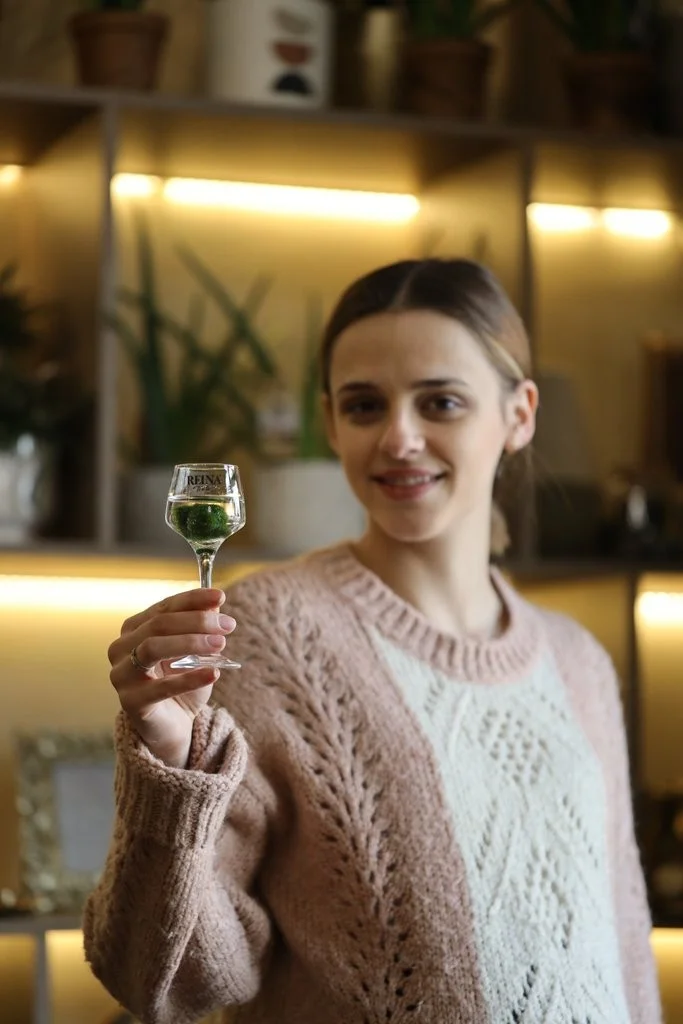 Osoba drži čašu u ruci u unutrašnjem prostoru s biljkama i zlatnim osvjetljenjem. Na čaši piše: "PELINKOVAC".