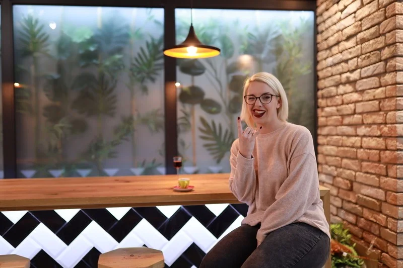 Osoba sjedi, pokazuje znak rukom, smije se, u modernom kafiću sa zidom od opeke i biljnom pozadinom. Na stolu su šoljica i čaša.