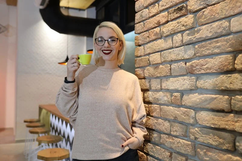 Osoba drži šoljicu, nasmijana, stojeći pored zida od cigle u kafiću.