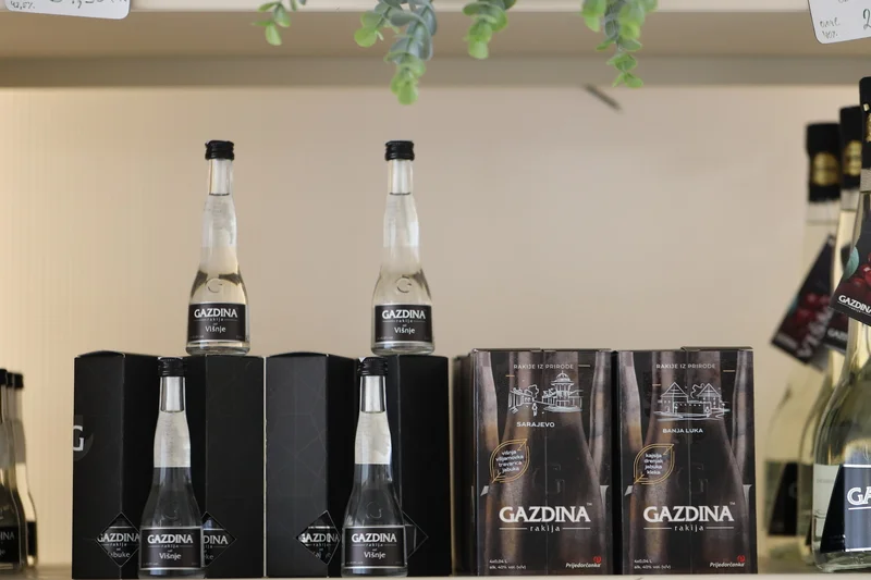 Boce i kutije s natpisom "GAZDINA rakija Višnje" stoje na polici, ukrašene visećim zelenim lišćem.