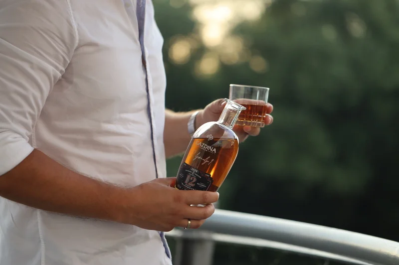 Osoba drži bocu pića i čašu, stoji na balkonu. Natpis na boci: "LAZDINA".
