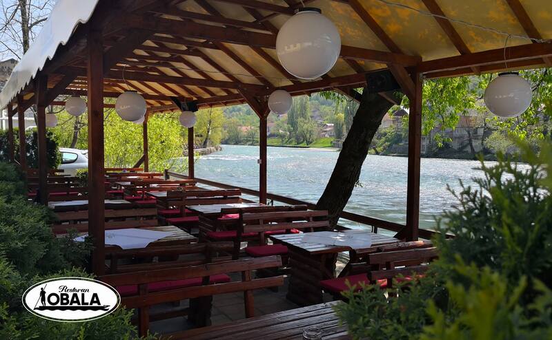 Drveni stolovi i klupe stoje na terasi pored rijeke, pod pokrivačem, okruženi zelenilom. Tekst: "Restoran OBALA".