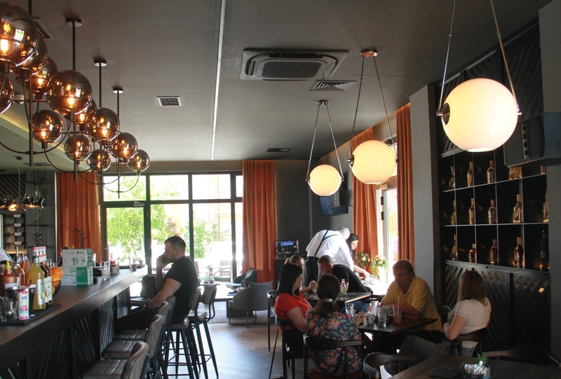 Ljudi sjede za stolovima, piju i razgovaraju u modernom kafiću s visećim rasvjetom i policama za piće.