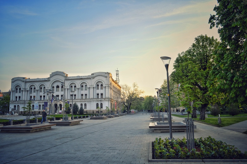 Zgrada stoji pored popločanog šetališta, okružena drvećem i klupama u urbanom parku tokom dana bez prisustva ljudi.