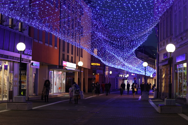 Ljudi hodaju ispod visećih plavih svjetala u večernjoj ulici. Znakovi: "Souvenirs", "Restoran".