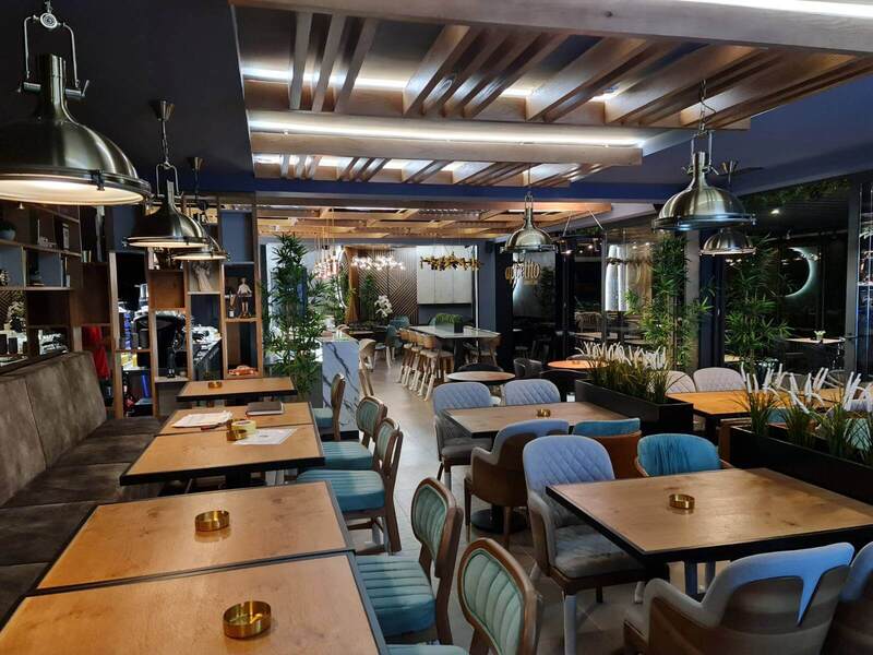 Stolovi i stolice stoje u restoranu sa drvenim detaljima, svjetiljkama i biljkama. Na zidu piše "appetito".