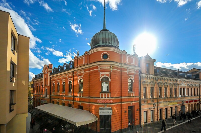 Zgrada crvene boje sa kupolom stoji na uglu ulice, natpis "RESTORAN CITADELA" vidljiv iznad ulaza, sunčano nebo u pozadini.