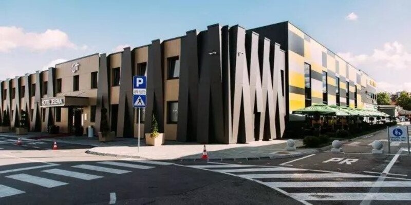 Zgrada hotela stoji pored ceste, okružena parking znakovima i zelenim suncobranima. Tekst: "HOTEL JELENA".