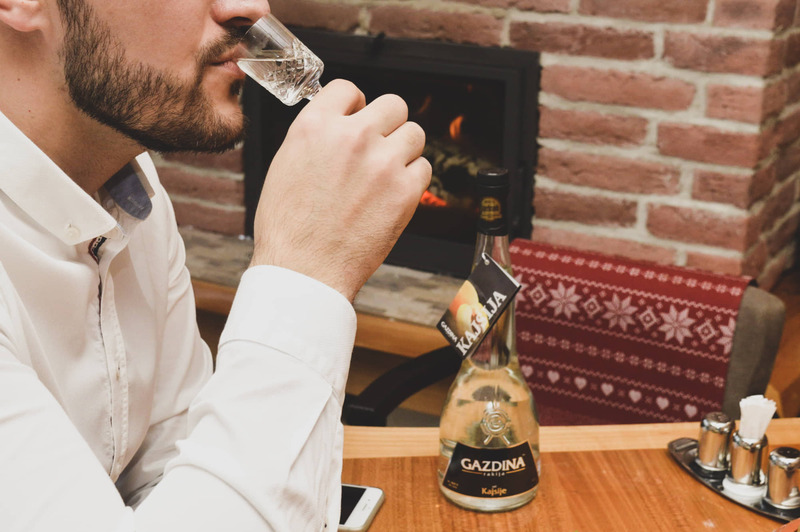 Čovjek pije rakiju u restoranu ispred kamina. Flaša označena "GAZDINAC Kašljača" i čaša na stolu.