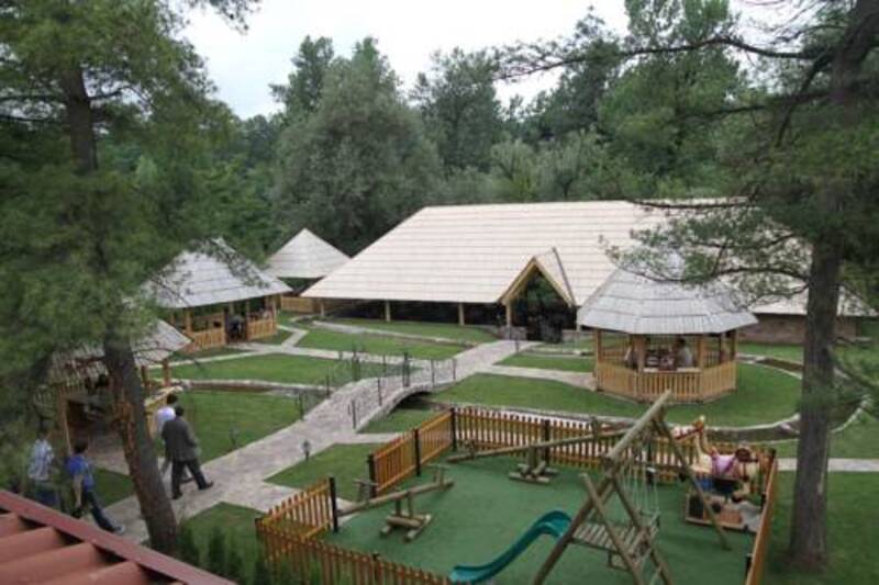 Drvena igrališta stoje mirno u parku okruženom stablima, dok ljudi šetaju stazama pored natkrivenih sjenica.