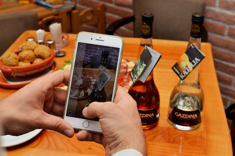 Ruke drže telefon, fotografišući flaše sa etiketom "GAZDINA" na drvenom stolu, u restoranskom okruženju.