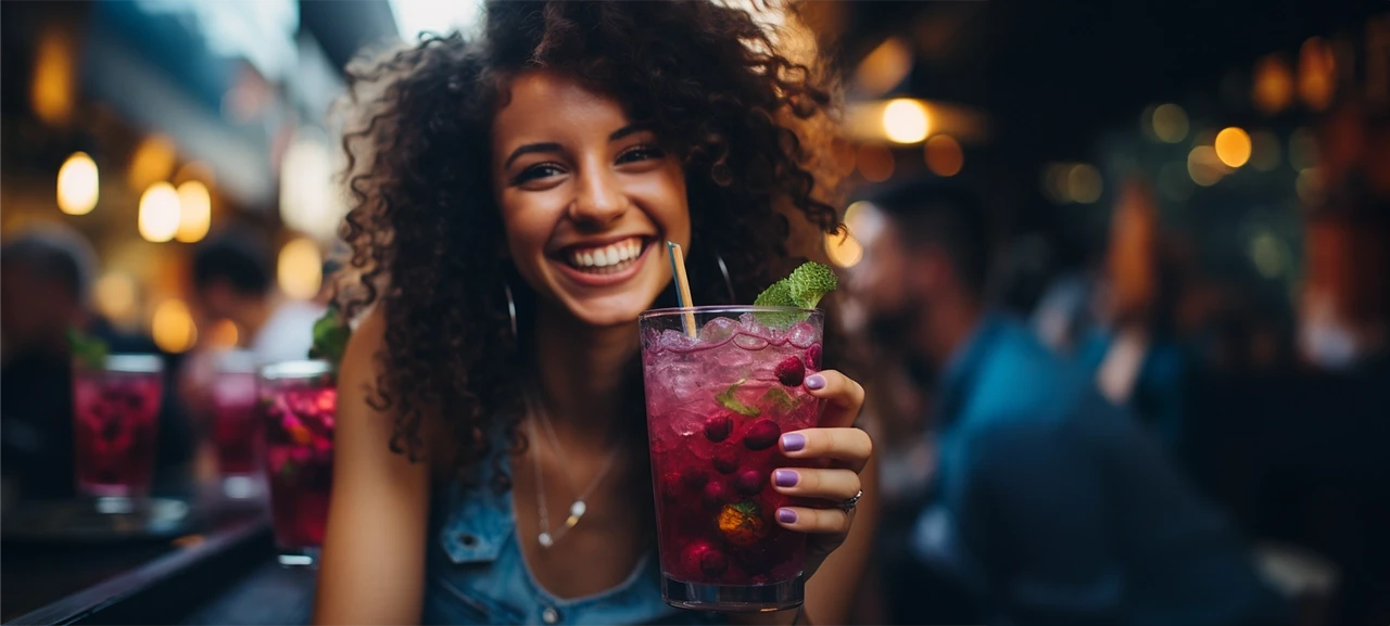 Osoba drži piće, smiješeći se u baru, okružena ljudima i prigušenim svjetlima.