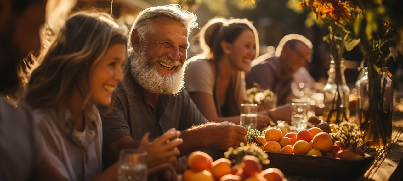 Ljudi se smiju za stolom prepunim voća, uživajući u druženju na otvorenom tokom sunčanog dana.