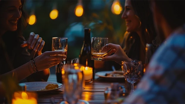 Ljudi nazdravljaju pićem za stolom, okruženom svijećama i toplim osvjetljenjem, tokom večernjeg okupljanja.